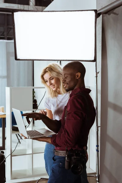 Photographe afro-américain et modèle caucasien choisissant des photos sur ordinateur portable ensemble pendant la séance photo en studio — Photo de stock