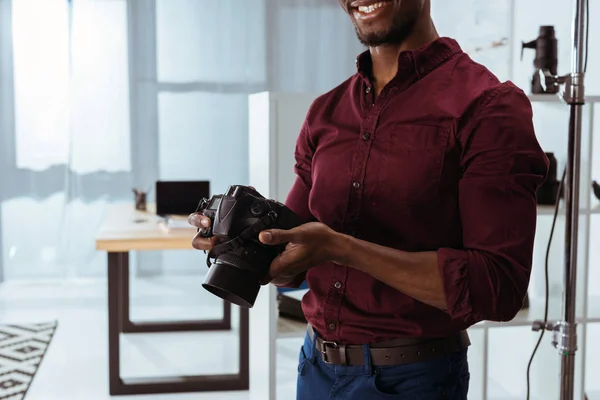 Tiro recortado de sorrindo fotógrafo afro-americano segurando câmera fotográfica em mãos em estúdio — Fotografia de Stock