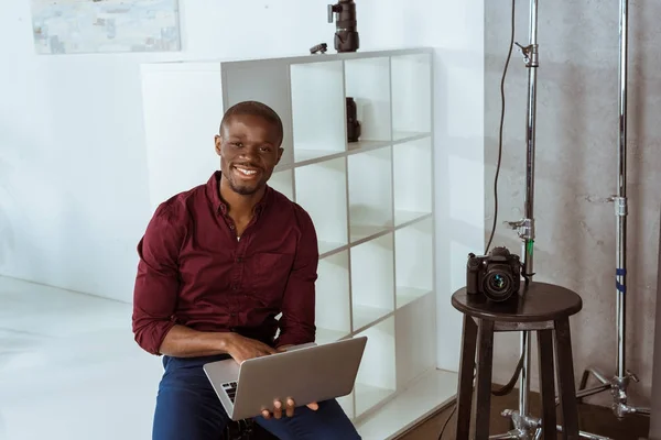 Portrait of cheerful african american photographer with laptop looking at camera in studio — Stock Photo