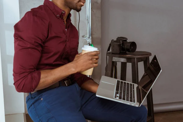 Cropped shot of african american photographer with coffee to go working on laptop in studio — Stock Photo