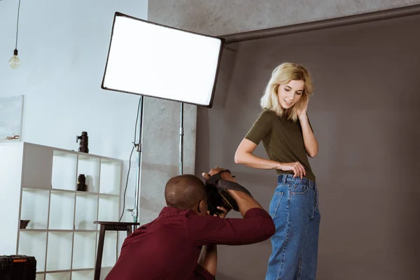 Beautiful caucasian woman posing while african american photographer taking picture in studio — Stock Photo