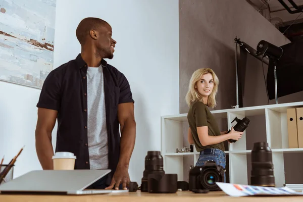 Fotógrafos multiculturales sonrientes mirándose antes de trabajar en el estudio - foto de stock