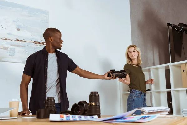 Smiling caucasian photographer giving photo camera to african american colleague at workplace in studio — Stock Photo