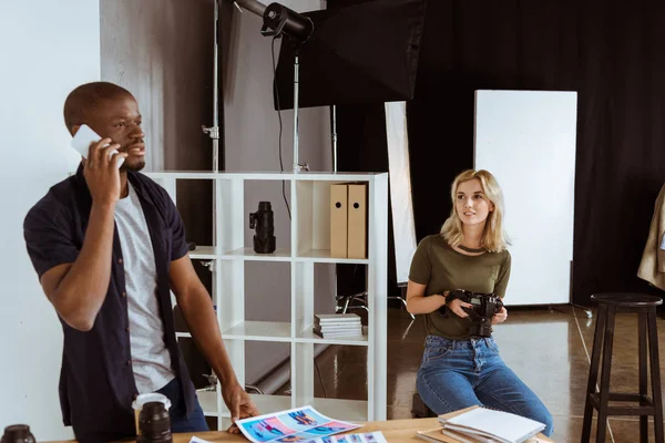 Afican american photographer talking on smartphone while caucaisan colleague choosing photos in studio — Stock Photo