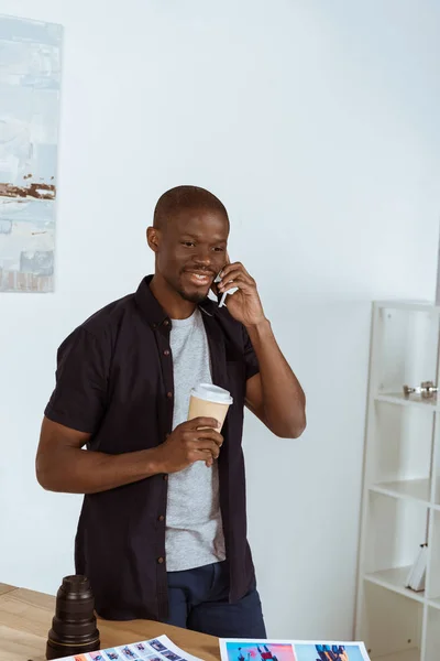 Portrait of smiling african american man with coffee to go talking on smartphone in office — Stock Photo