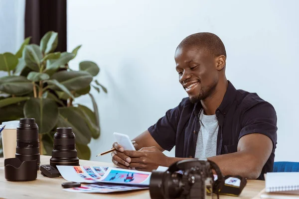 Portrait d'un homme afro-américain souriant utilisant un smartphone alors qu'il était assis sur le lieu de travail avec un équipement de photographie — Photo de stock