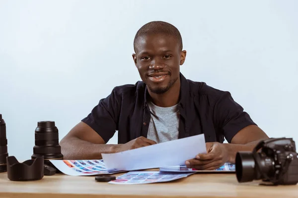 Portrait of smiling african american photographer sitting at workplace with photoshoot examples and photo camera and looking at camera — Stock Photo