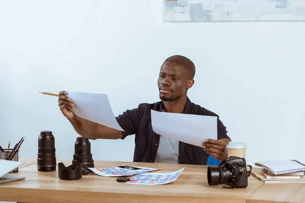 Retrato de fotógrafo afroamericano enfocado mirando ejemplos de sesión de fotos en el lugar de trabajo - foto de stock