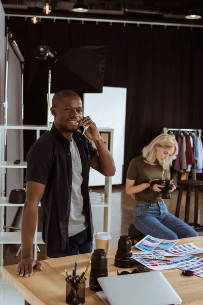 Fotógrafo americano afican hablando en el teléfono inteligente mientras colega caucaso elegir fotos en el estudio - foto de stock