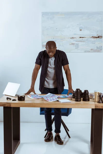 Fotógrafo afroamericano concentrado mirando ejemplos de sesión de fotos mientras está parado en el lugar de trabajo - foto de stock
