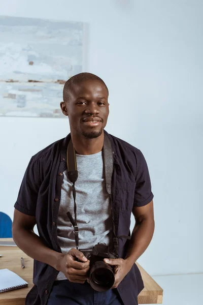 Portrait of african american photographer with photo camera leaning on workplace in office — Stock Photo