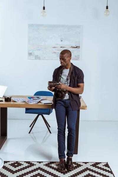 Африканский американский фотограф с фотокамерой, опирающийся на рабочее место в офисе — стоковое фото