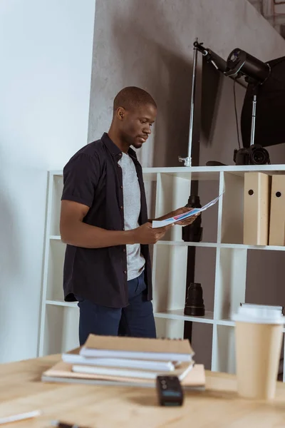 Side view of focused african american photographer looking at papers in hands in studio — Stock Photo