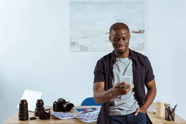 Retrato de un fotógrafo afroamericano sonriente usando un teléfono inteligente mientras se apoya en la mesa con equipo de fotografía en la oficina - foto de stock