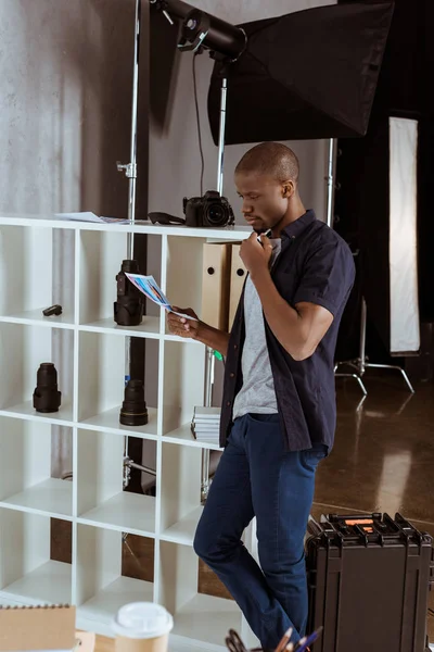 Vista lateral do fotógrafo afro-americano focado olhando para papéis na mão em estúdio — Fotografia de Stock