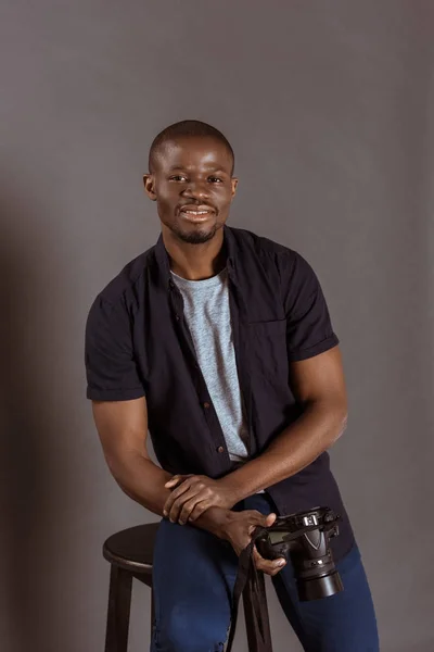 Retrato de sonriente fotógrafo afroamericano con cámara de fotos sentado en la silla y mirando a la cámara - foto de stock