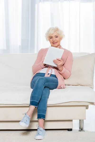 Joyeuse femme âgée assise sur le canapé et utilisant une tablette numérique à la maison — Photo de stock