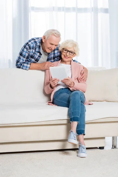 Senior umarmt Ehefrau mit digitalem Tablet in der Hand auf Sofa zu Hause — Stockfoto