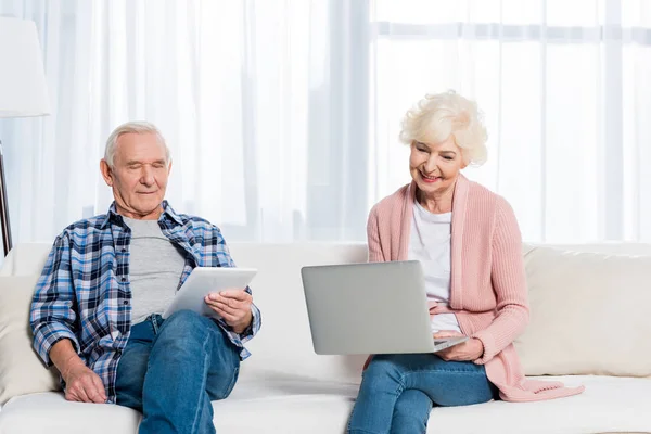 Retrato de la sonriente esposa y esposo mayores usando dispositivos digitales en casa - foto de stock
