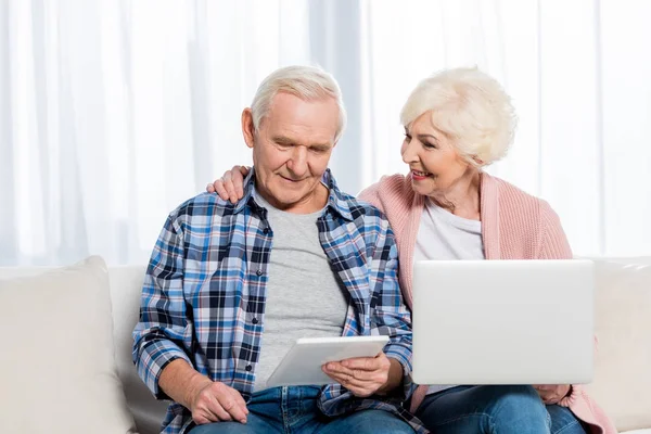 Retrato de la sonriente esposa y esposo mayores usando dispositivos digitales en casa - foto de stock