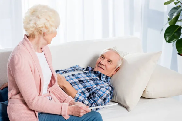 Homme âgé souriant regardant femme avec tablette dans les mains tout en se reposant sur le canapé à la maison — Photo de stock