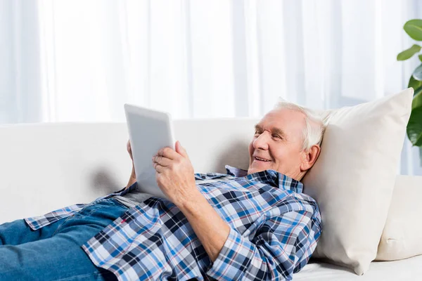 Side view of cheerful senior man lying on sofa and using tablet at home — Stock Photo