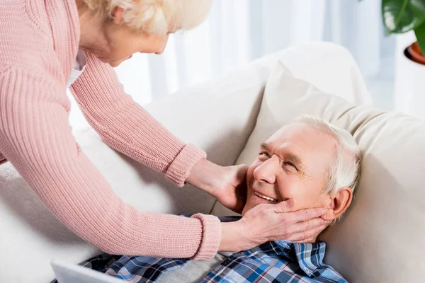Старшая женщина осторожно прикасается к мужу, пока он лежит на диване — стоковое фото
