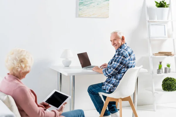 Vista parcial del hombre mayor en la mesa con el ordenador portátil mirando a la esposa sentada en el sofá en casa - foto de stock