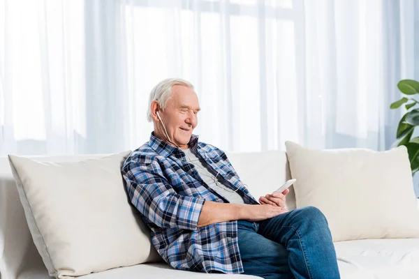 Vista lateral de sorrir homem idoso em fones de ouvido com smartphone descansando no sofá em casa — Fotografia de Stock