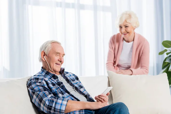 Selective focus of senior man in earphones listening music while wife standing near by at home — Stock Photo