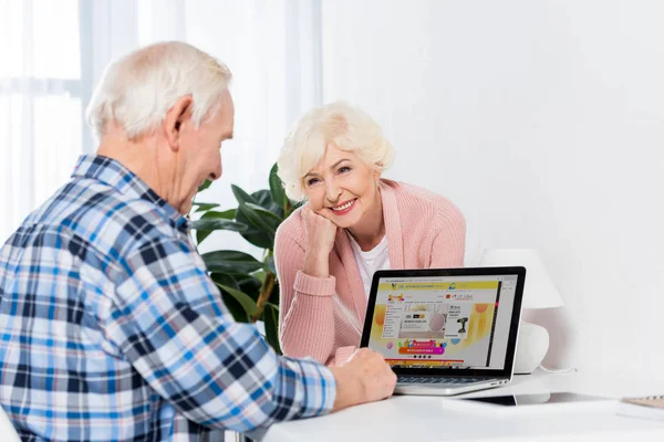 Porträt einer Seniorin, die ihren Mann zu Hause mit Laptop mit aliexpress-Logo betrachtet — Stockfoto