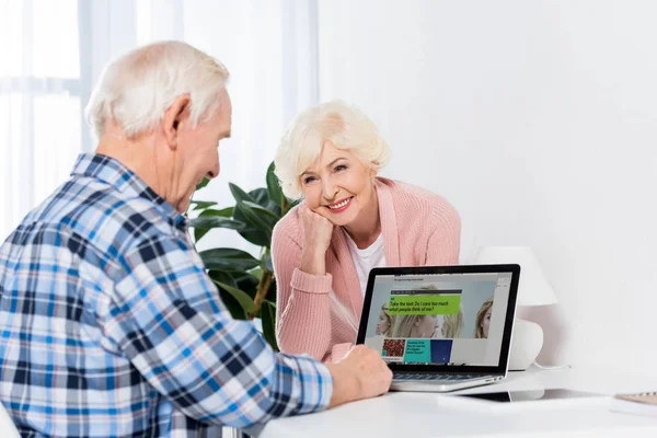 Retrato de la esposa mayor mirando al marido usando el ordenador portátil con el logotipo de la penes en casa - foto de stock