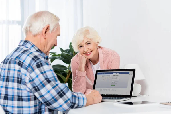 Portrait of senior woman looking at husband working on laptop with facebook logo at home — Stock Photo