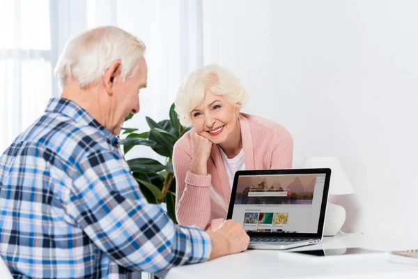 Retrato de la mujer mayor feliz mirando al marido usando el ordenador portátil en casa - foto de stock