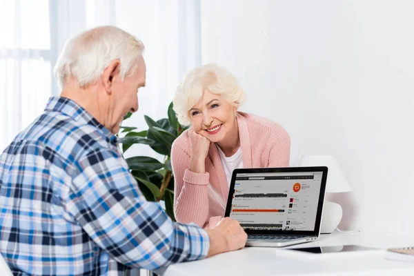 Retrato de la mujer mayor mirando al marido que trabaja en el ordenador portátil con el logotipo de soundcloud en casa - foto de stock