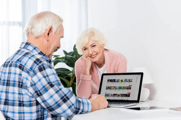 Ritratto di donna anziana allegra che guarda il marito usando il computer portatile a casa — Foto stock