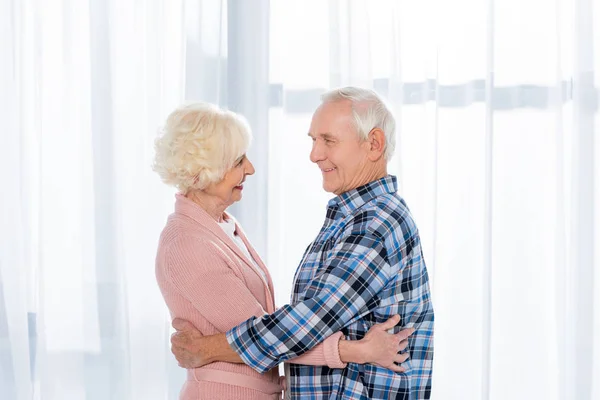 Vista lateral de alegre pareja mayor abrazándose y mirándose el uno al otro - foto de stock