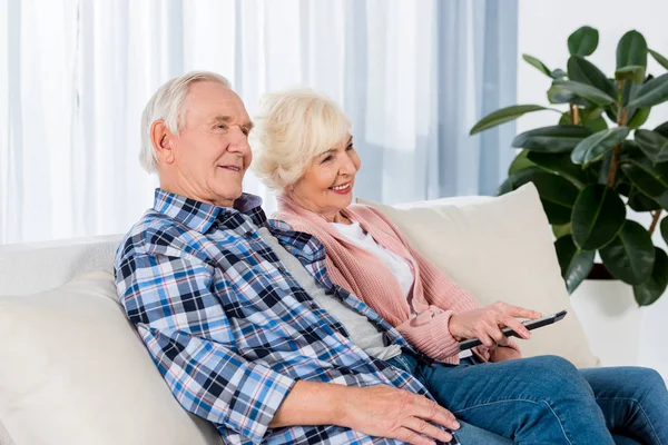 Feliz pareja de ancianos con control remoto viendo la televisión en el sofá - foto de stock
