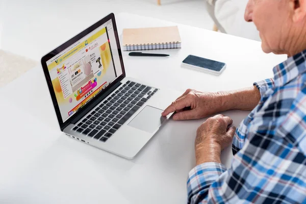 Partial view of senior man sitting at table and using laptop with aliexpress logo — Stock Photo