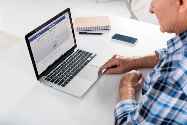 Teilansicht eines älteren Mannes bei der Arbeit am Laptop mit Facebook-Logo am Tisch mit Notebook zu Hause — Stockfoto