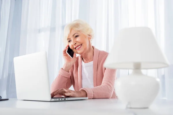 Portrait de femme âgée souriante parlant sur smartphone tout en utilisant un ordinateur portable à la maison — Photo de stock