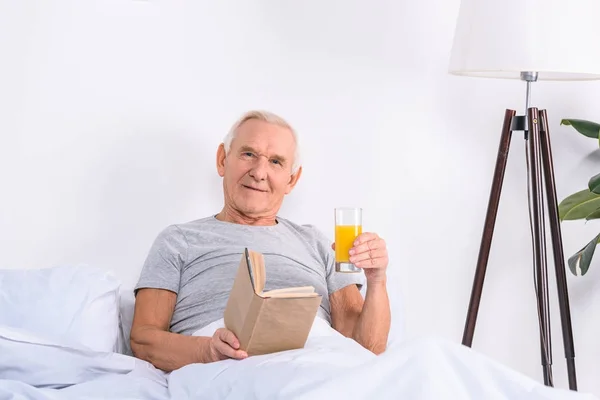 Retrato de hombre mayor con vaso de jugo y libro en las manos mirando a la cámara mientras está acostado en la cama en casa - foto de stock
