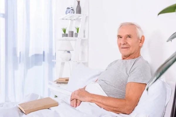 Vue latérale de l'homme âgé avec livre reposant dans le lit et regardant la caméra à la maison — Photo de stock