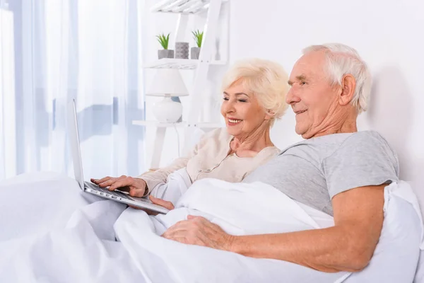 Pareja de ancianos utilizando el ordenador portátil mientras descansa en la cama en casa - foto de stock