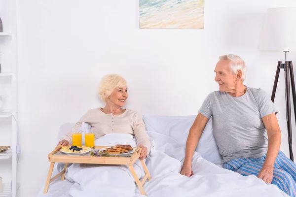 Mulher sênior alegre com café da manhã na cama olhando para o marido perto de em casa — Fotografia de Stock