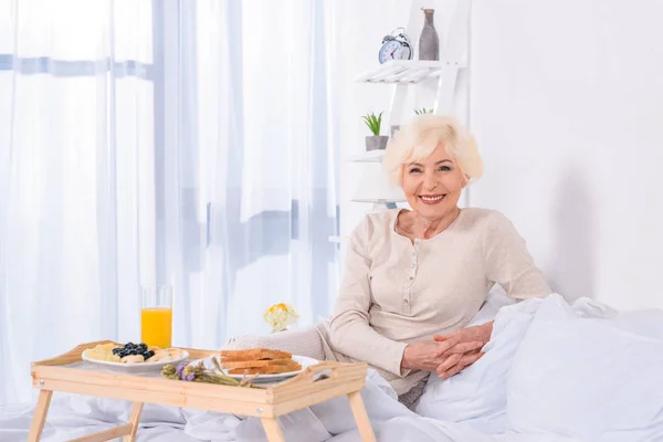 Ritratto di donna anziana felice con colazione a letto guardando la macchina fotografica a casa — Foto stock