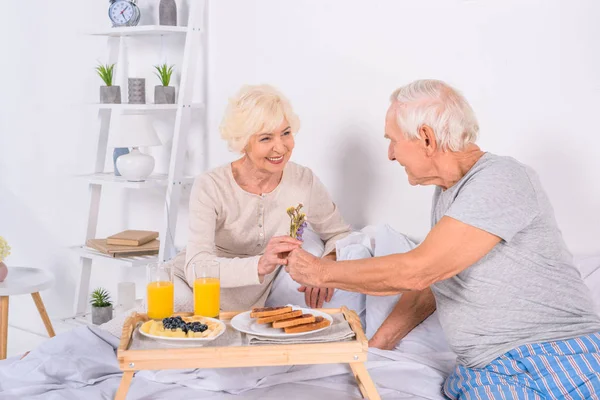 Uomo anziano che presenta fiori alla moglie mentre fa colazione a letto insieme a casa — Foto stock