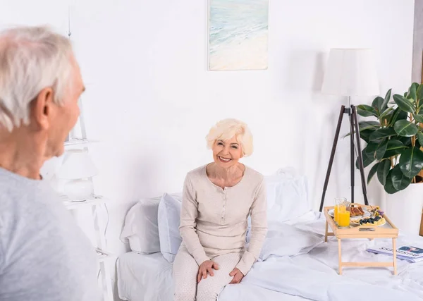 Happy senior woman sitting on bed and looking at husband at home — Stock Photo