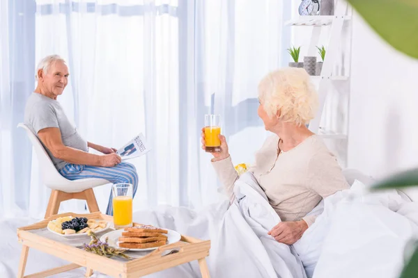 Side view of senior woman having breakfast in bed while husband wit newspaper sitting at table  in bedroom — Stock Photo