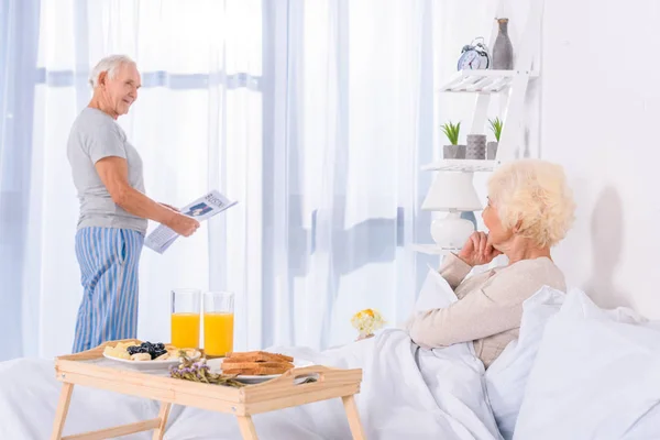 Teilansicht der Seniorin, die Ehemann mit Zeitung am heimischen Fenster ansieht — Stockfoto